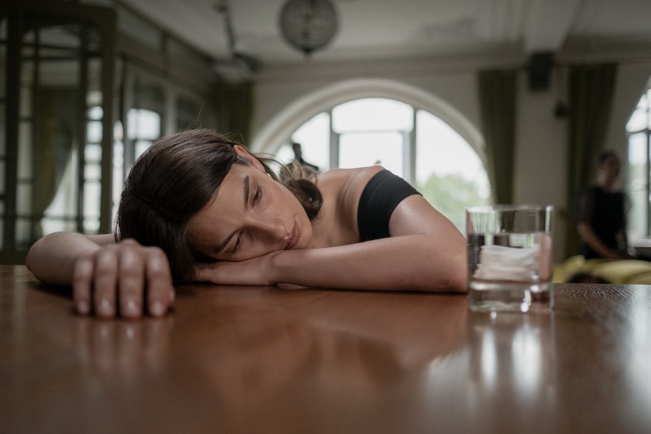 portfolio-img-05 A woman in a black dress rests her head on a table beside a glass of water, conveying sadness.
