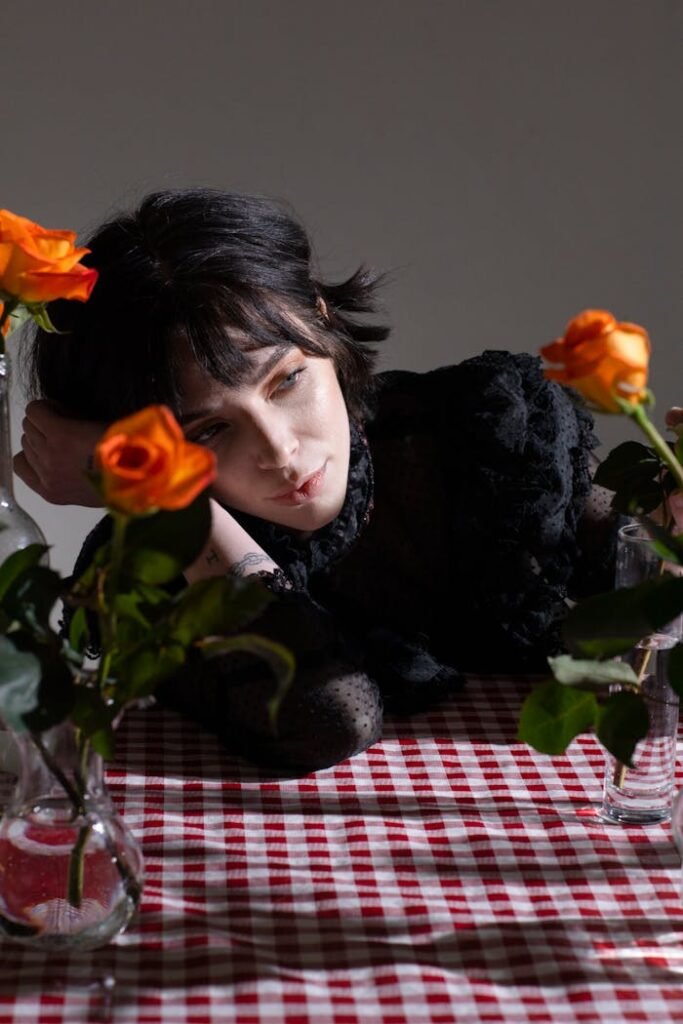 A thoughtful woman leans on a table adorned with vibrant orange roses.